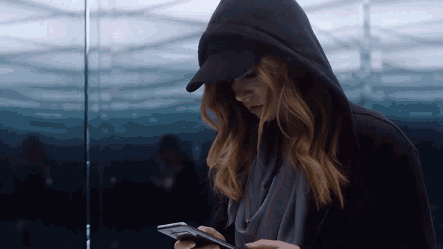 a woman wearing a hooded jacket and a hat looks at her phone
