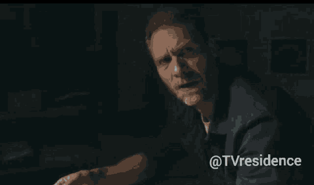 a man with a beard is sitting in a dark room with the words @tvresidence behind him