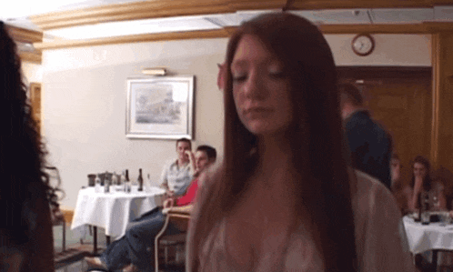 a woman with long red hair is standing in a room with people sitting at tables