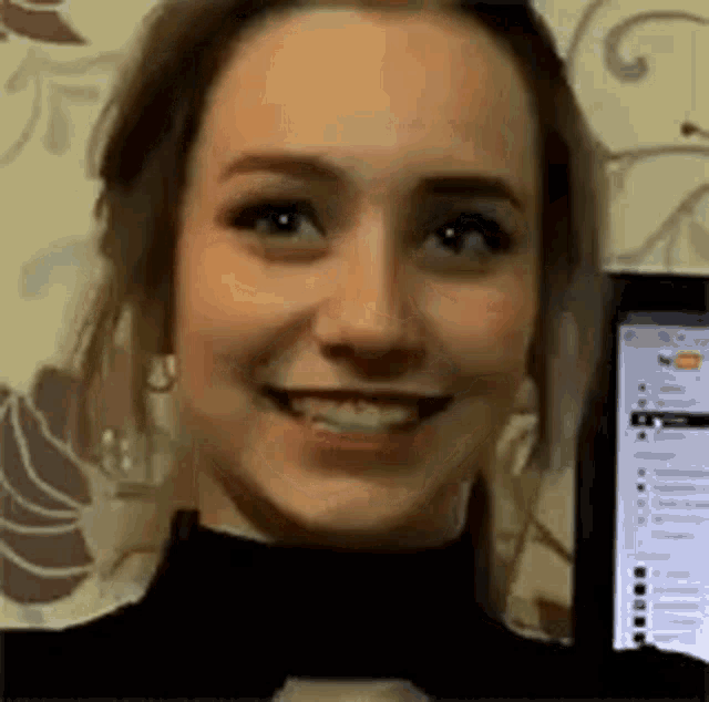 a woman is smiling while holding a cell phone in front of a computer screen .