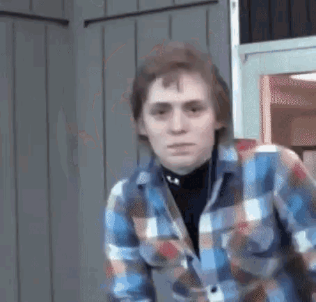 a young man wearing a plaid shirt and a black turtleneck is standing in front of a door .