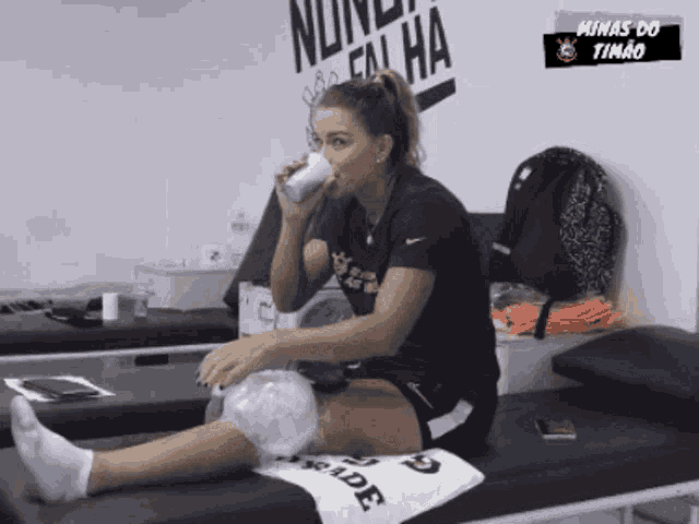 a woman sits on a bed drinking from a cup with minas do timão written on the wall
