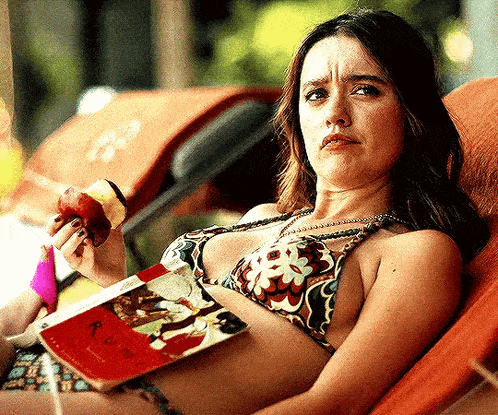 a woman in a bikini is eating an apple and reading a book while laying in a chair .