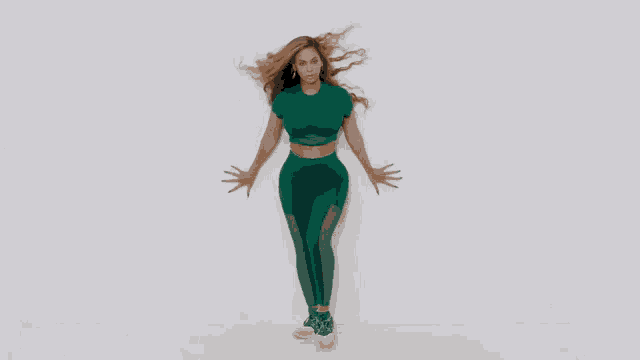 a woman in a green crop top and leggings is standing in front of a white wall with her arms outstretched .