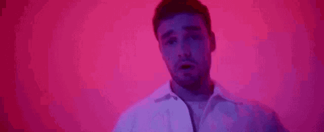 a man in a white shirt is standing in front of a pink wall and looking at the camera .