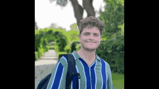 a young man wearing a striped shirt and carrying a backpack is standing in a park .
