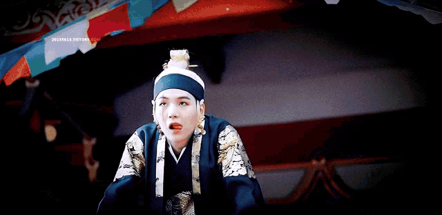 a man in a blue and gold costume with a bun on his head stands in front of a colorful flag