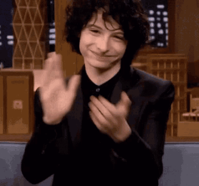 a young man in a suit and black shirt is clapping his hands and smiling .