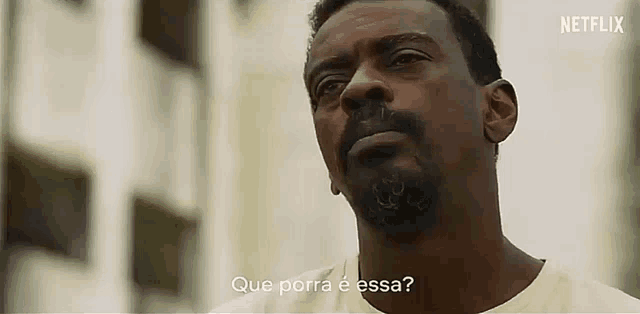 a man with a beard is standing in front of a building with the words que porra e essa written on the bottom .