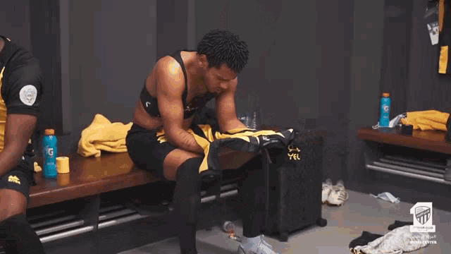 a man sits on a bench in a locker room next to a yellow suitcase that says yel