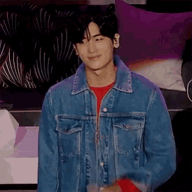 a young man wearing a denim jacket and a red sweater is standing in front of a couch .