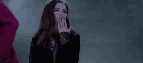 a woman in a black jacket is standing in front of a wall with her hand up .