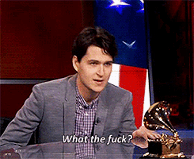 a man in a suit and plaid shirt is sitting at a table and says " what the fuck "