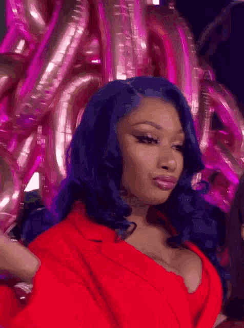 a woman with blue hair is wearing a red top and standing in front of pink balloons .