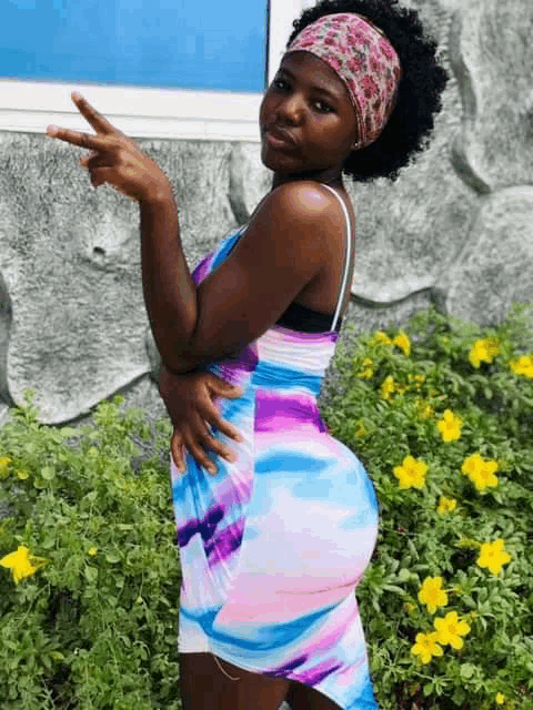 a woman wearing a headband and a colorful dress is standing in front of flowers and giving a peace sign .