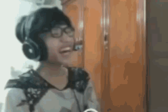a woman wearing headphones and glasses is laughing while standing in front of a wooden cabinet .