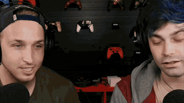 two men wearing headphones are talking into microphones in front of a wall with video game controllers on it .