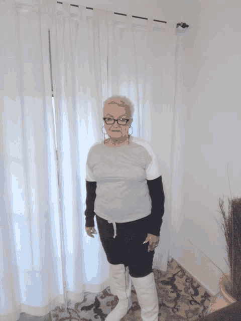 an older woman wearing glasses and white boots stands in front of a white curtain