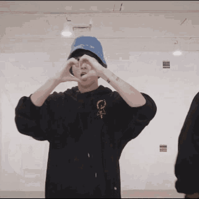 a man wearing a black hoodie and a blue hat is making a heart with his hands