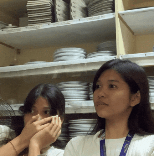 two girls covering their mouths in front of a shelf with plates