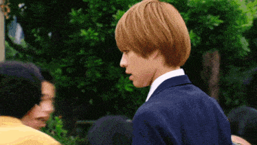a young man in a suit and white shirt stands in front of trees