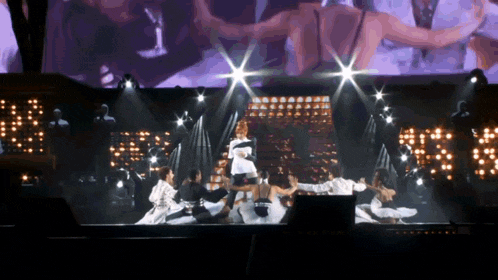 a group of people dancing on a stage with a large screen behind them