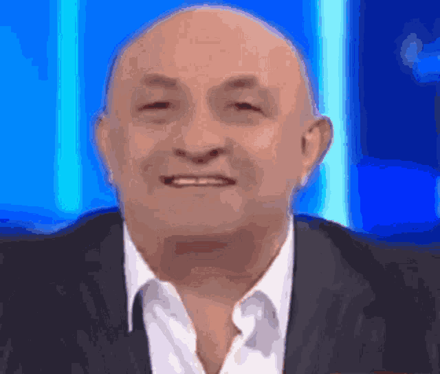 a bald man wearing a suit and a white shirt is smiling in front of a blue background .
