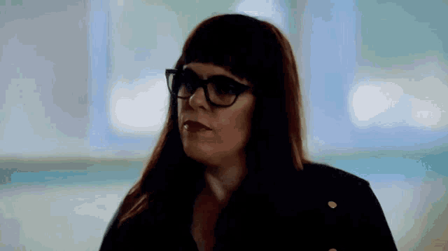 a woman wearing glasses and a black jacket looks to the side