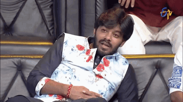 a man in a floral shirt is sitting on a couch with a tv channel behind him