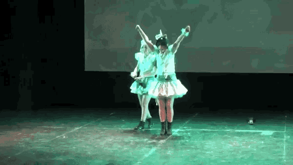 two women are dancing on a stage with their arms in the air and a green light behind them .