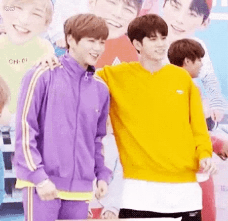 two young men are standing next to each other in front of a poster . one of the men is wearing a yellow sweater .