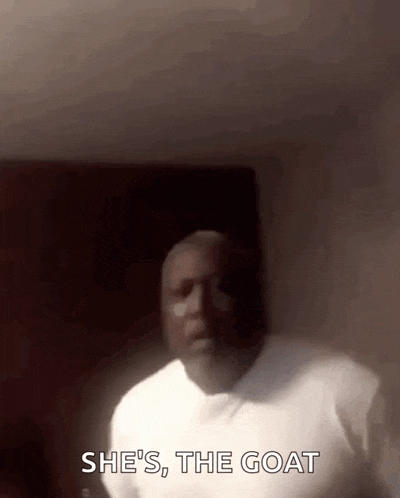 a man in a white shirt is standing in a dark room and saying she 's the goat .
