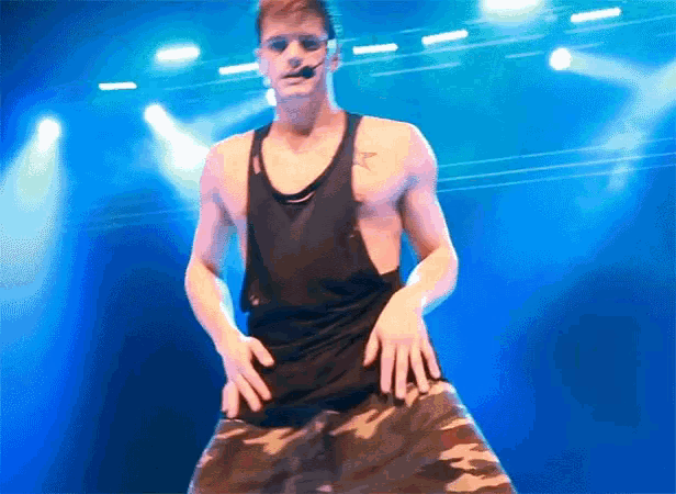 a shirtless man in a black tank top and camo shorts is dancing on a stage