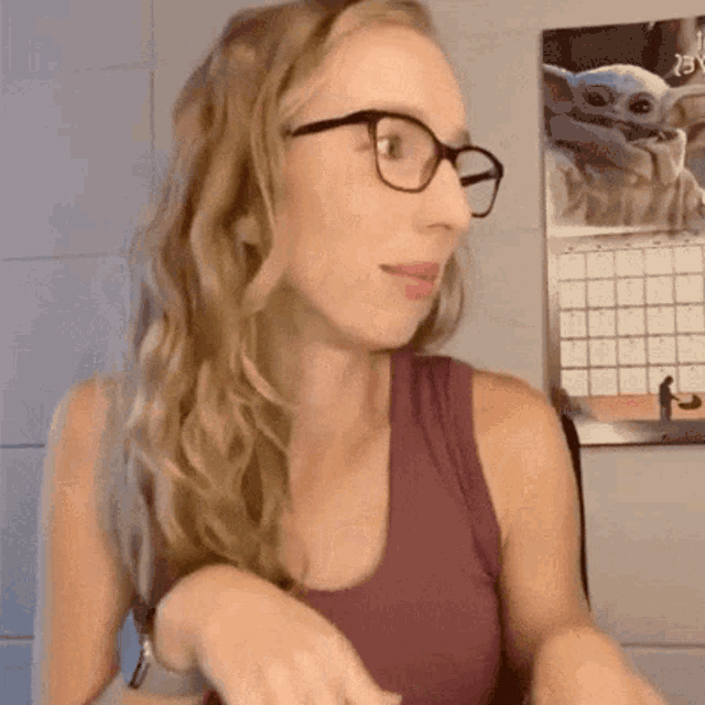 a woman wearing glasses and a purple tank top is standing in front of a wall with a calendar on it .