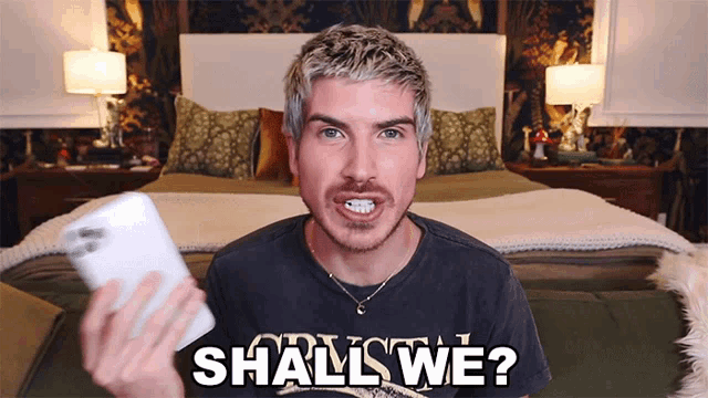 a man holding a cell phone with the words " shall we " on his shirt