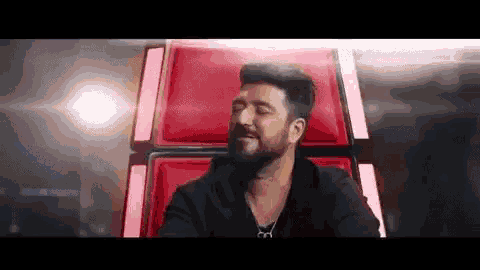 a man with a beard is sitting in a red chair with his eyes closed and his mouth open .