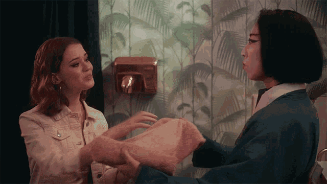 two women are talking in front of a wall with palm leaves on it
