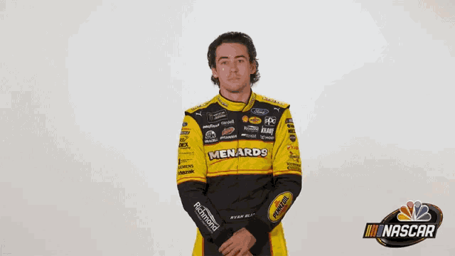 a man wearing a yellow and black menards racing suit is standing with his arms outstretched .