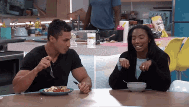 a man and a woman are sitting at a table eating food with a bag of baked beans behind them