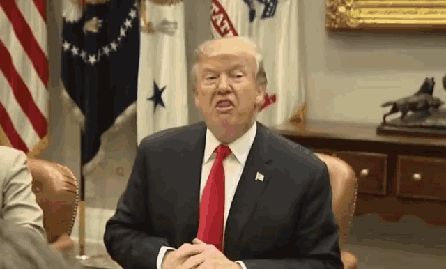 donald trump is sitting at a desk in front of american flags and making a face