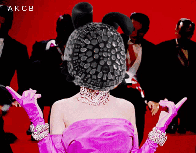 a woman in a pink dress and gloves stands in front of a group of men and the word akcb is on the bottom