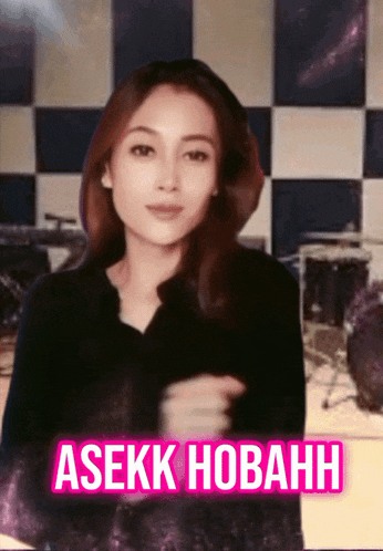 a woman in a black shirt is standing in front of a checkered wall with the words asekk hobahn in pink letters