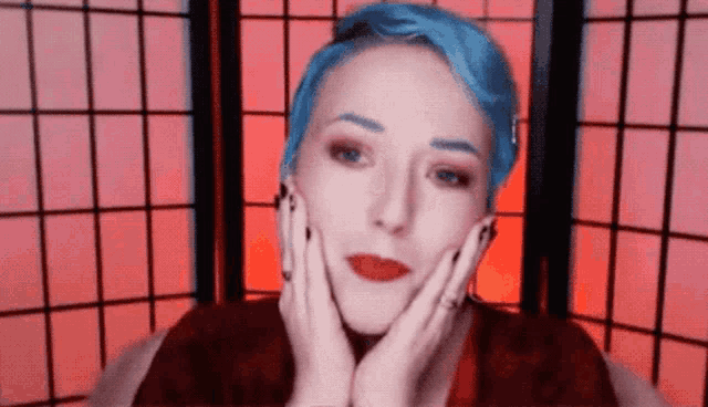 a woman with blue hair has her hands on her face in front of a red screen