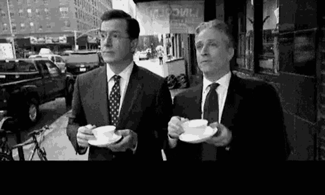 two men in suits and ties are standing next to each other on a city street holding cups of coffee .