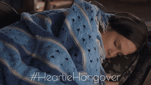 a woman wrapped in a blue blanket is sleeping on a couch with #heartiehangover written below her