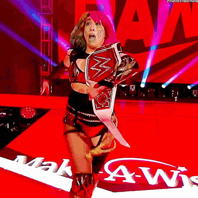 a female wrestler with pink hair is holding a championship belt in front of a sign that says mah-a-wis