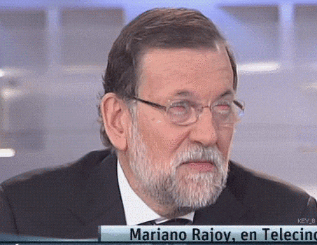 a man with glasses and a beard is sitting in front of a sign that says mariano rajoy