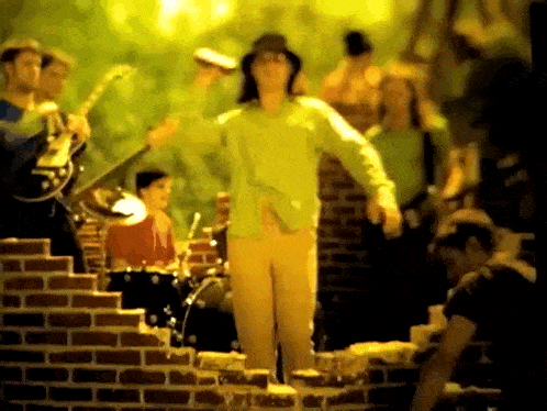 a man in a green shirt is standing in front of a brick wall with a band behind him .