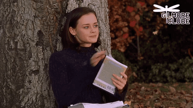 a woman leaning against a tree reading a book with gilmore club on the bottom