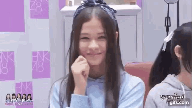 a girl wearing a blue shirt and a headband is smiling with her hands on her chin .